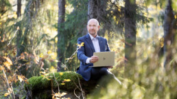 Mies istuu puku päällä ja kannettava tietokone sylissään metsässä, auringonvalo pilkahtelee lehtien läpi