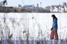 Tumma hiuksinen nainen kävelee lumen peittämällä järven jäällä, jota reunustaa kaislikko.
