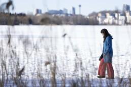 Tumma hiuksinen nainen kävelee lumen peittämällä järven jäällä, jota reunustaa kaislikko