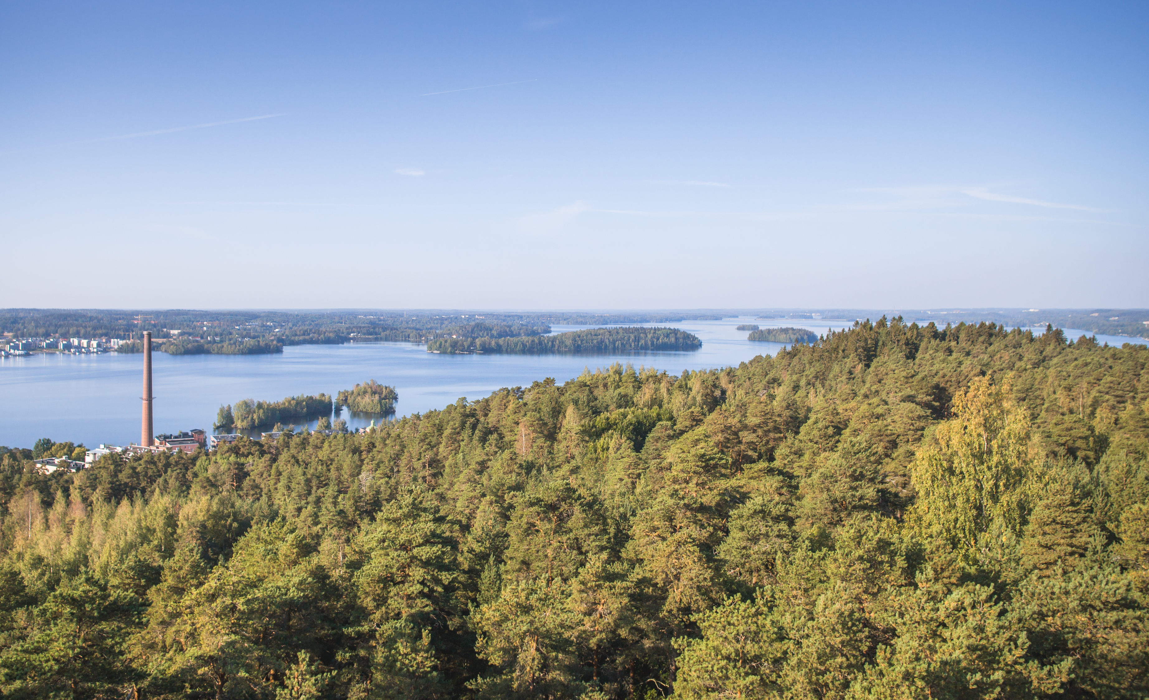 Maisemakuva Tampereesta lintuperspektiivistä, metsän takana siintää sinisenä kimmeltävä Pyhäjärvi.
